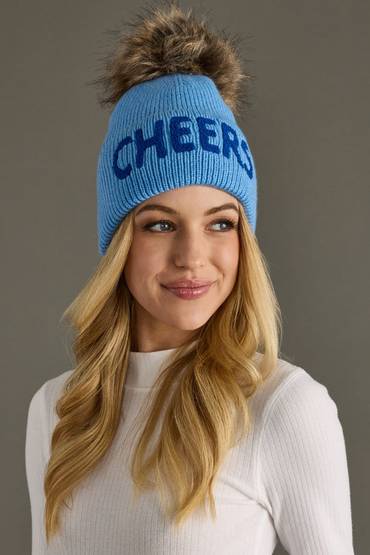 Woman wearing a blue knit beanie with 'CHEERS' text against a gray background