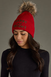 A woman with long dark hair wears the Panache Apparel Collection Merry & Bright Pom Hat - Red & Green, paired with a black ribbed top. She looks down slightly with a soft smile.
