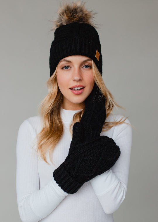 Woman wearing a black knit beanie with a fur pom-pom and matching gloves against a gray background