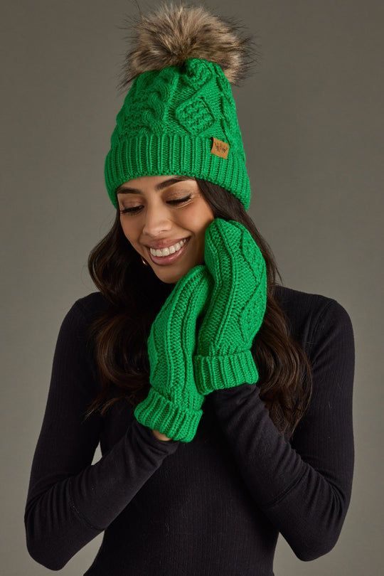 Person wearing a green knit hat and gloves with a brown pom-pom on a gray background