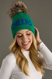 Person wearing a green beanie with 'CHEERS' text and fur pom-pom on a gray background