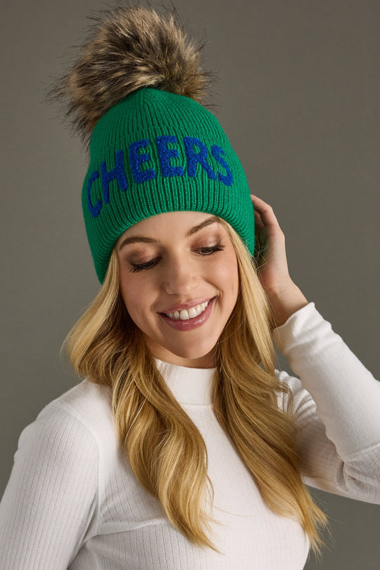 Person wearing a green beanie with 'CHEERS' text and fur pom-pom on a gray background