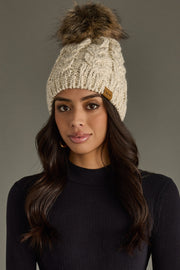 Woman wearing a beige knitted beanie with a fur pom-pom on a gray background