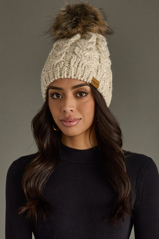Woman wearing a beige knitted beanie with a fur pom-pom on a gray background