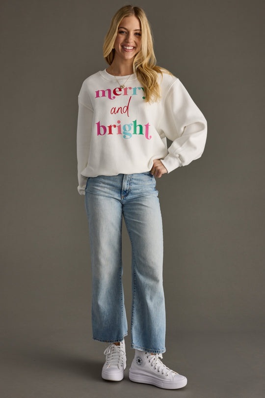A smiling woman poses against a gray background wearing the Panache Apparel Collection's Merry & Bright Sweatshirt in white, styled with light blue jeans and white high-top sneakers.