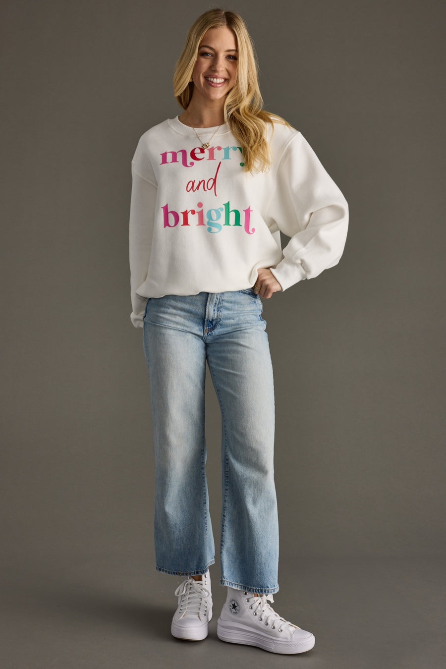 A smiling woman poses against a gray background wearing the Panache Apparel Collection's Merry & Bright Sweatshirt in white, styled with light blue jeans and white high-top sneakers.