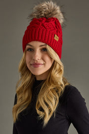 A woman with long blonde hair, dressed in a black ribbed turtleneck sweater, smiles and faces slightly to the right while wearing the Panache Apparel Collection Red Cable Knit Pom Hat, featuring a fleece-lined interior and a faux fur pom, against a gray background.