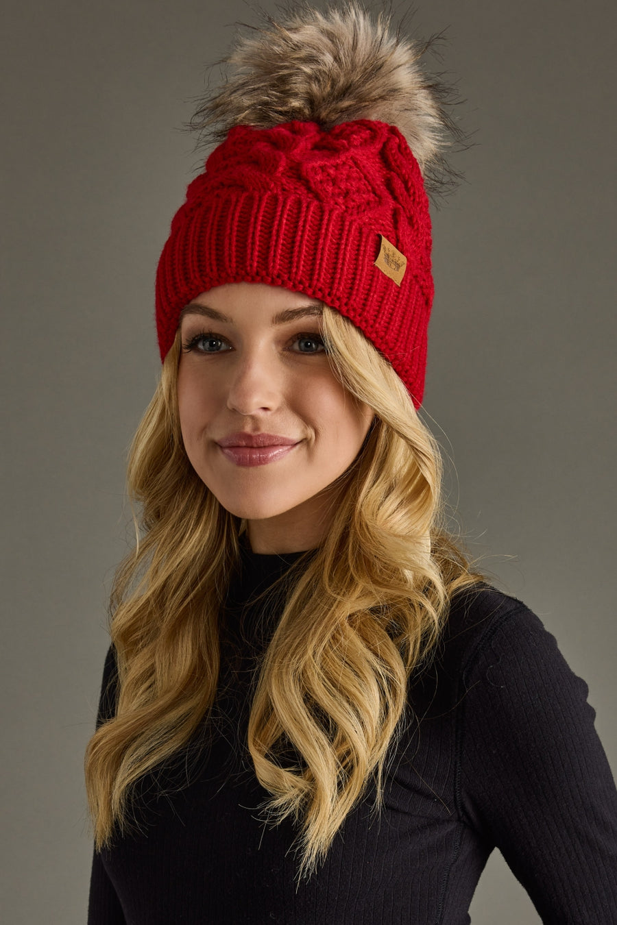 A woman with long blonde hair, dressed in a black ribbed turtleneck sweater, smiles and faces slightly to the right while wearing the Panache Apparel Collection Red Cable Knit Pom Hat, featuring a fleece-lined interior and a faux fur pom, against a gray background.