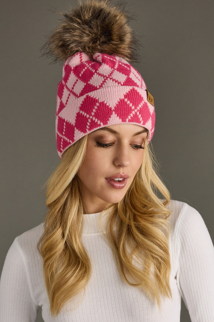 Woman wearing a pink and blush argyle beanie with a fur pom-pom on a gray background
